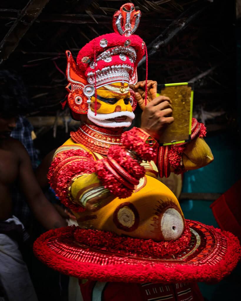 A candid, close-up shot of a Theyyam performer in the final stages of preparation. His face is heavily painted in traditional patterns of orange, yellow, and black. He holds a small, hand-held mirror, intently checking his reflection to ensure the sacred makeup is perfect. The lighting is soft and atmospheric, highlighting the focus in his eyes and the fine detail of the ancestral designs.