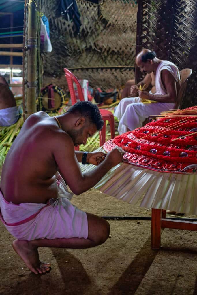 A vertical, documentary-style photograph of a craftsman crouching in a dimly lit preparation shed, meticulously working on a large, ornate Theyyam headdress. The headdress features vibrant red fabric and white pleated palm leaves. In the background, another man in traditional white attire is seen assisting with the preparations. The atmosphere is focused and industrious, highlighting the manual craftsmanship behind the festival.