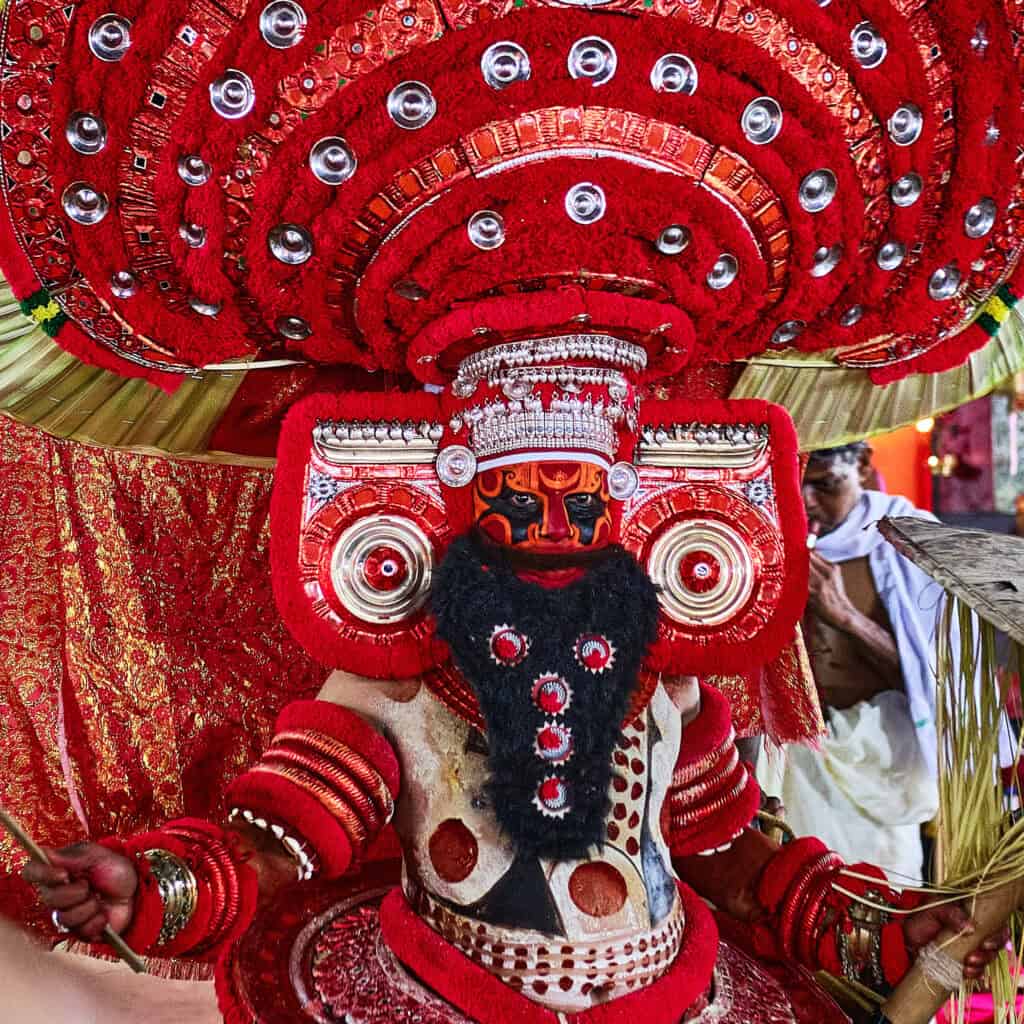 A square, front-facing portrait of a Theyyam performer in full ceremonial regalia. He wears a massive, circular red headdress adorned with silver reflective discs and an intricate red and black facial mask with a prominent black beard. His eyes look directly into the lens with intense focus. The background shows a hint of the ceremonial setting and another person in traditional white attire, emphasizing the vibrant red of the deity's costume