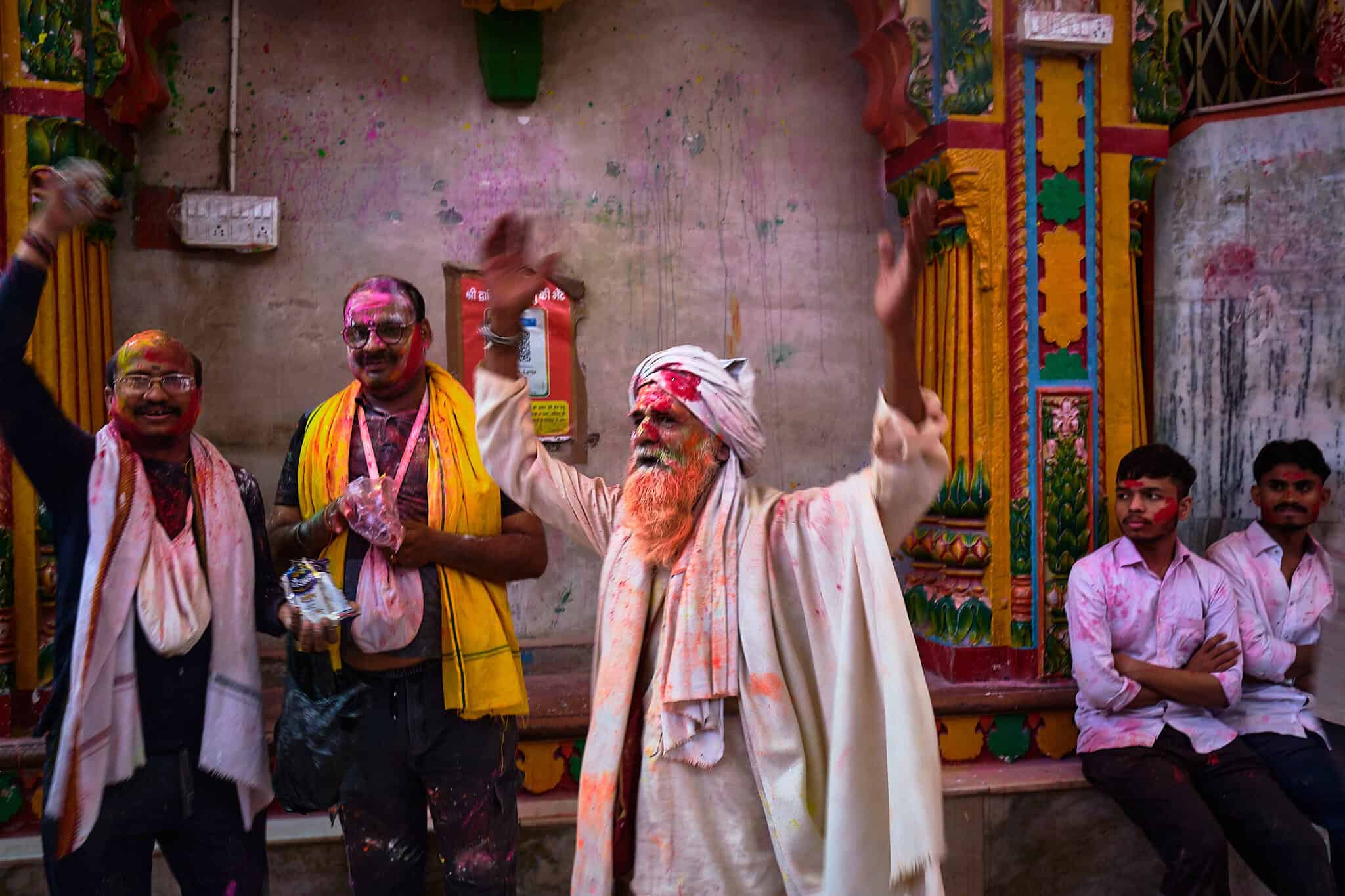 A vibrant morning scene inside the Dwarkadhish Temple in Mathura during Holi. A man in a white turban and shawl stands in the center with his arms raised in celebration, his beard and face dusted with orange powder. Around him, other men smile, their faces and clothes covered in pink and yellow pigment, against the backdrop of intricately carved and colored temple columns.