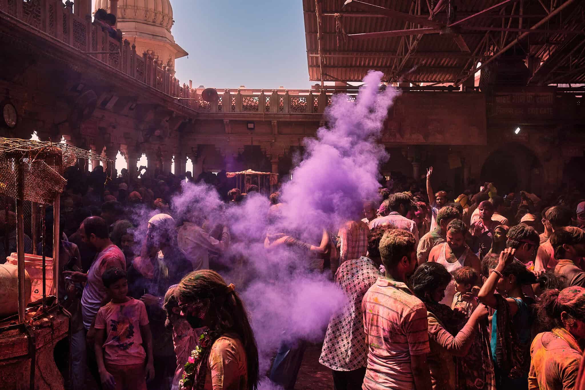 A wide shot of a temple courtyard filled with a large, towering cloud of purple and violet Holi powder smoke. The crowd below is bathed in the cool-toned light of the dust.
