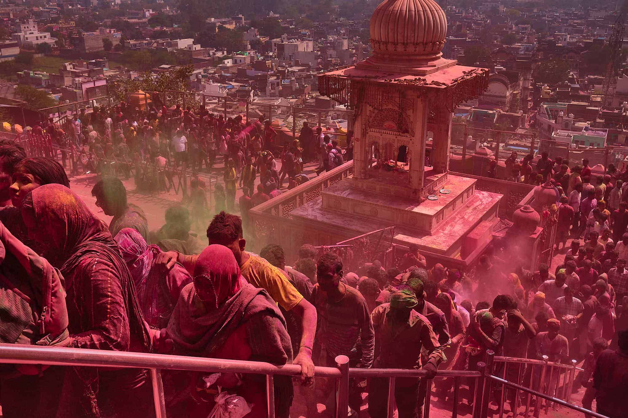 A high-angle, expansive shot from a temple staircase in Barsana during Holi. A thick, pink cloud of powder hangs in the air, tinting the entire scene—from the thousands of people packed onto the terraces to the urban sprawl of the city in the background—in a monochromatic magenta hue. In the foreground, people grip the silver handrails as they navigate the crowded stairs.