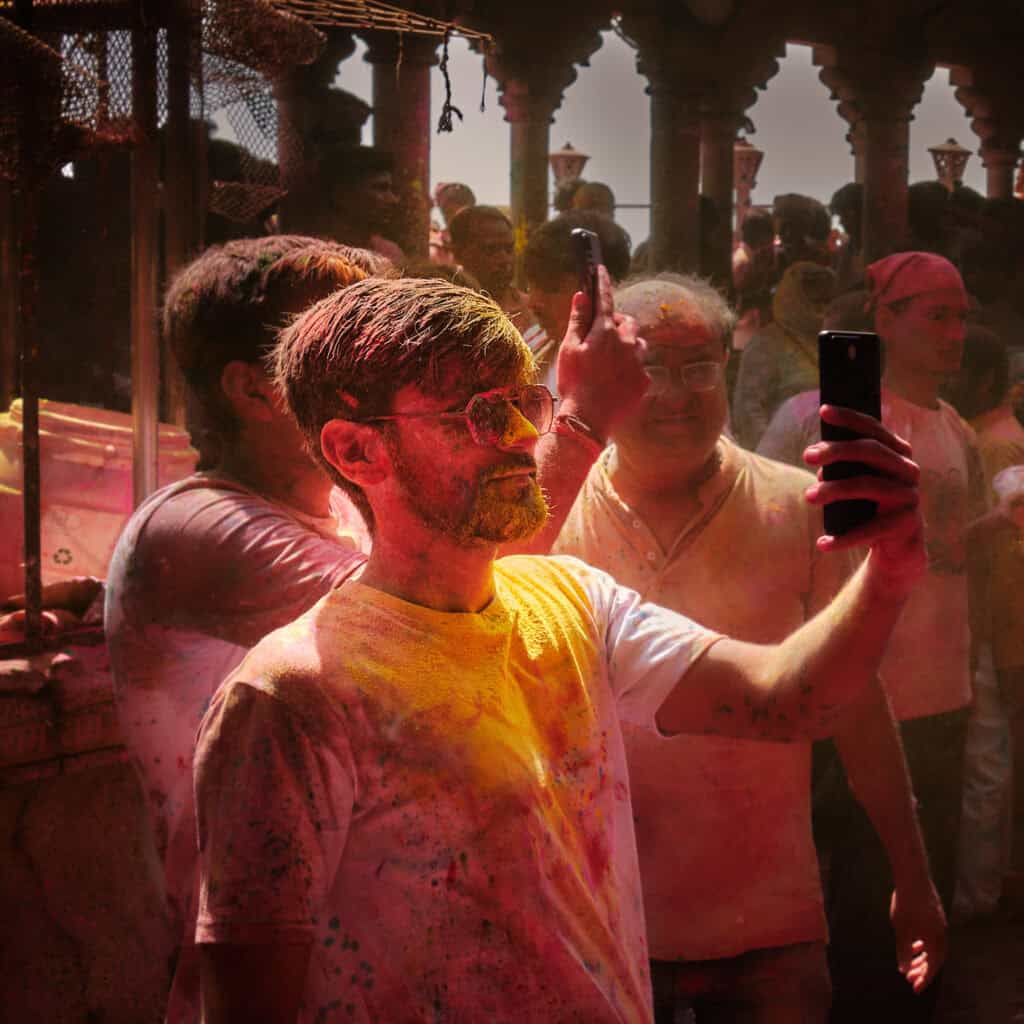 A person looking at a mobile phone while celebrating Holi, with colors visible on their face and clothing.