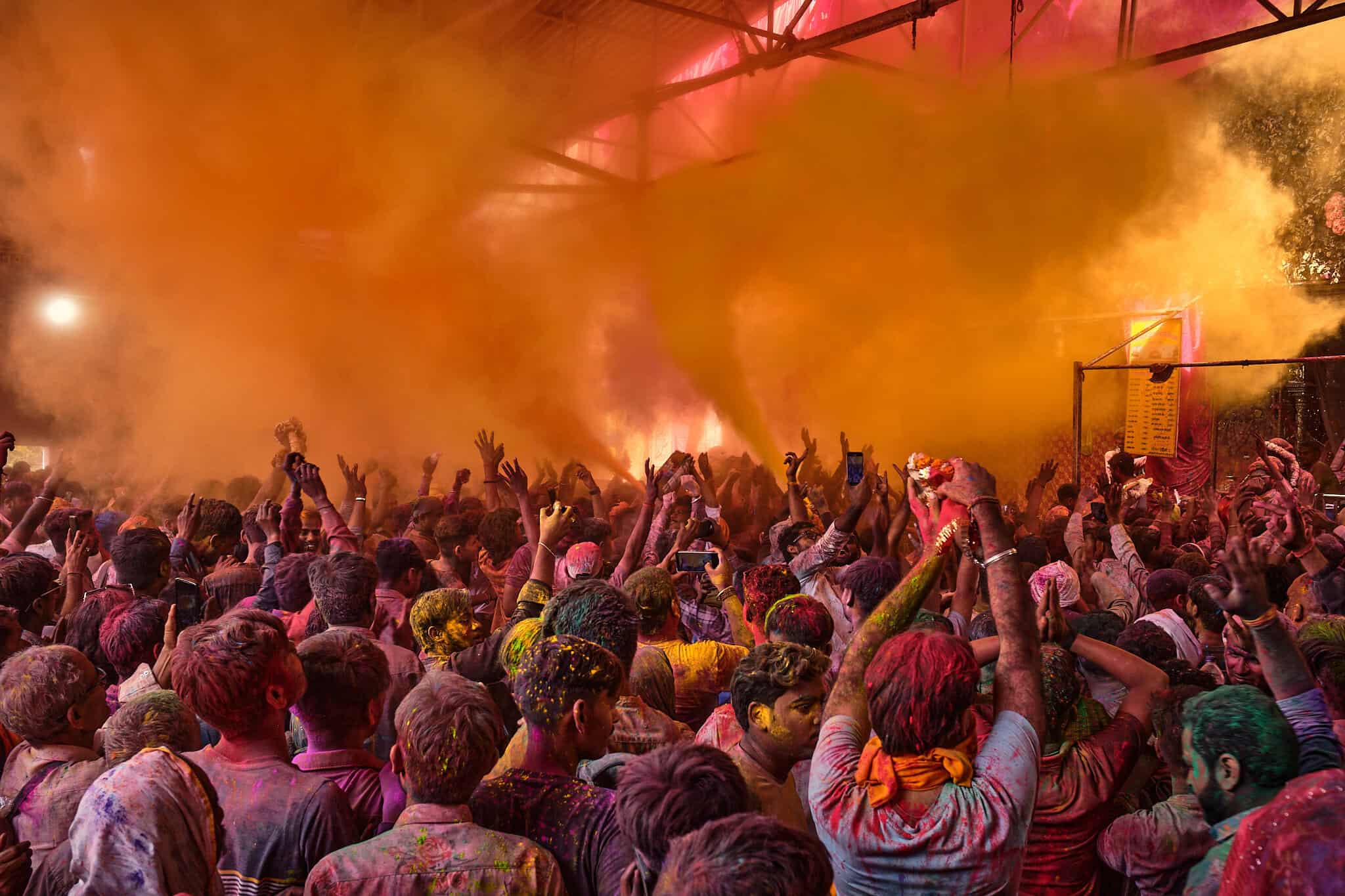 A high-energy shot of a crowd with their hands raised toward a thick, swirling plume of bright orange and yellow powder being sprayed through the air.