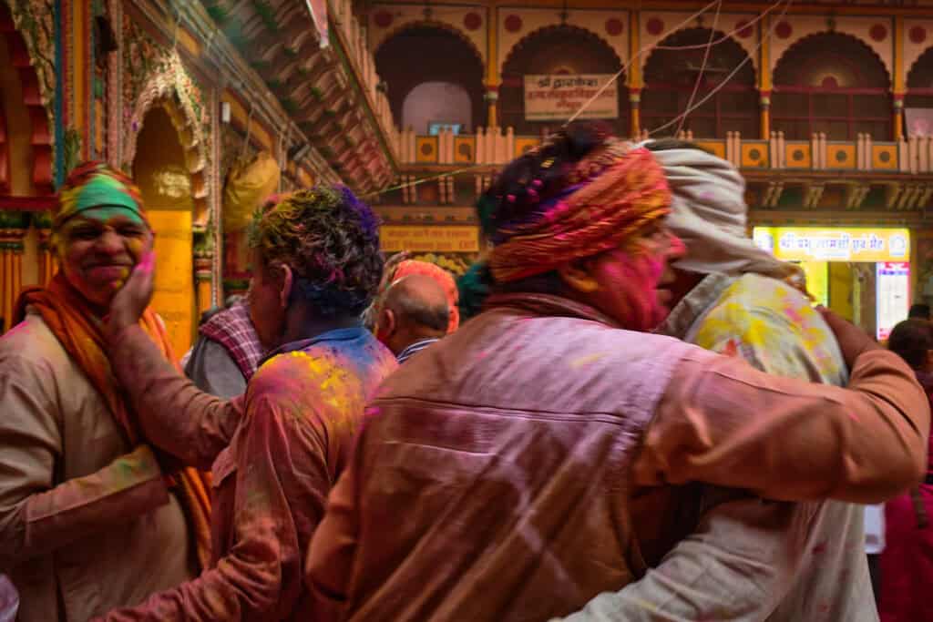 A candid, warm-toned photograph inside a Mathura temple during Holi. In the foreground, two men in pigment-stained clothes are locked in a friendly embrace, while to the left, another man smiles broadly as someone applies green and orange powder to his face. The background features the intricate, multi-layered balconies of the temple architecture.