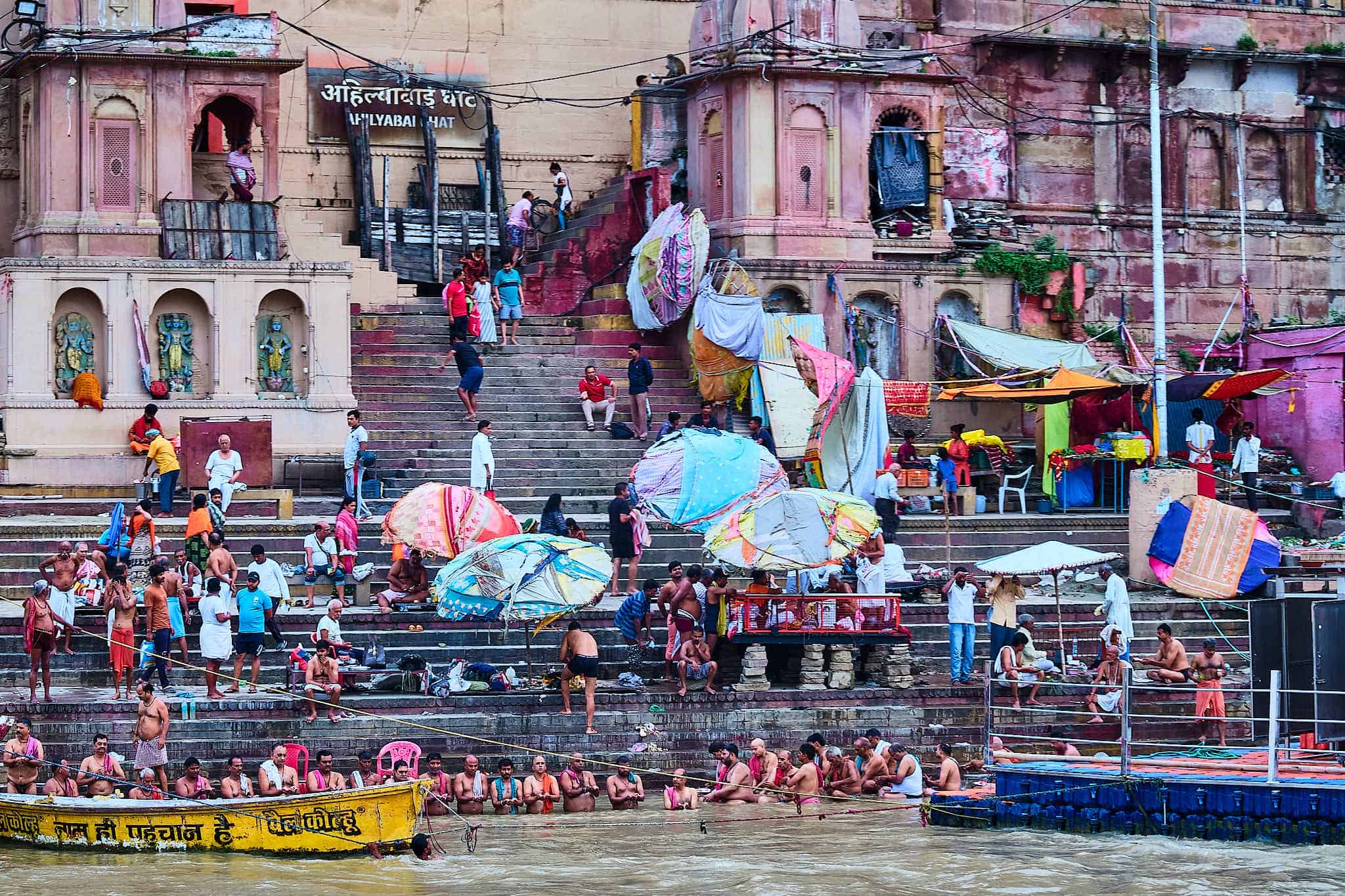 A high-angle, wide-angle shot of Ahilyabai Ghat in Varanasi, showing the stone steps crowded with people, colorful sun umbrellas, and pilgrims performing ritual baths in the Ganges in the foreground, with ancient buildings rising in the background.