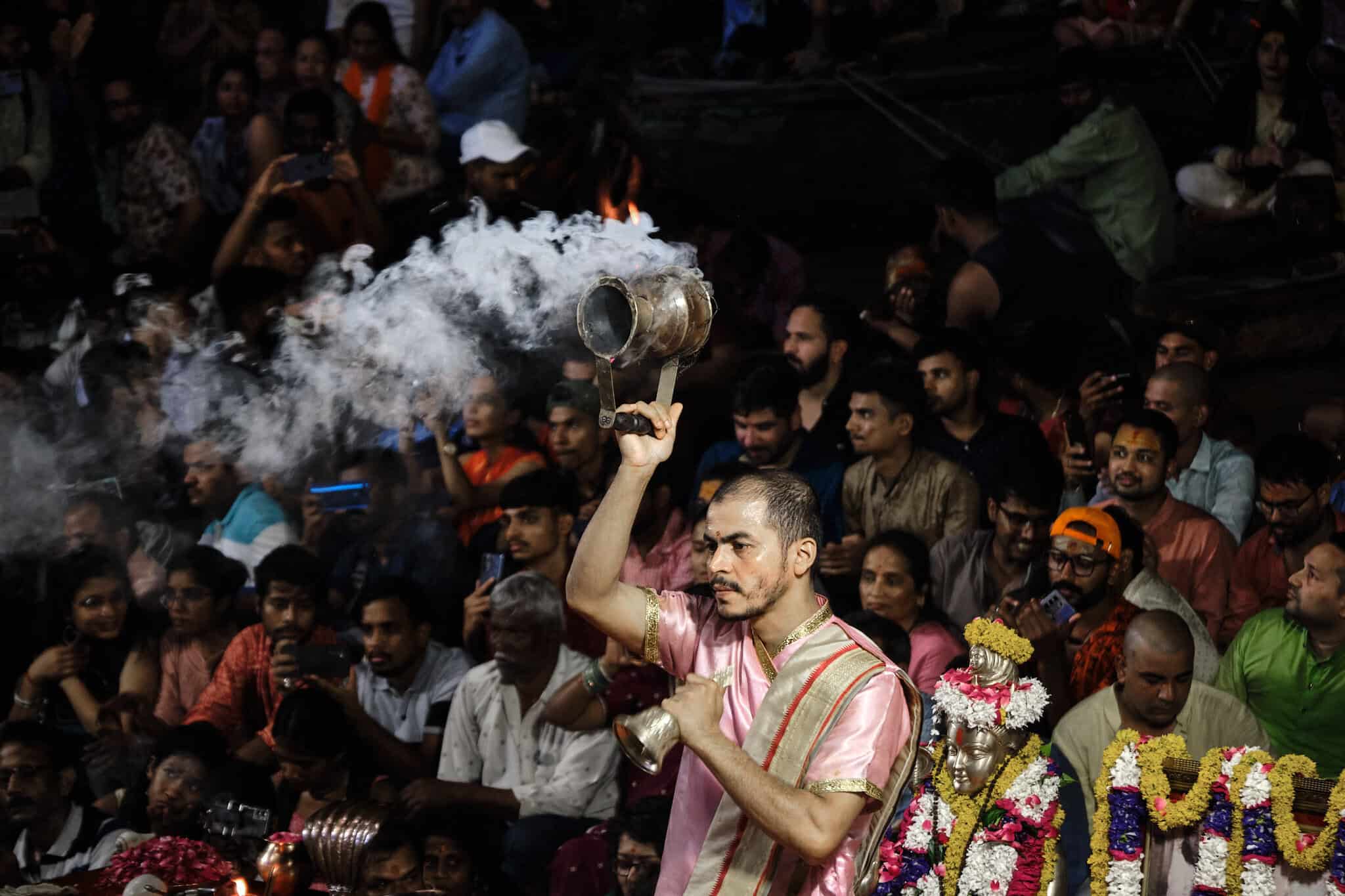 A medium shot of a young priest in a pink silk robe holding a smoking incense burner aloft during the evening Ganga Aarti. A large, diverse crowd of onlookers is visible in the background, many holding up smartphones to capture the ritual.
