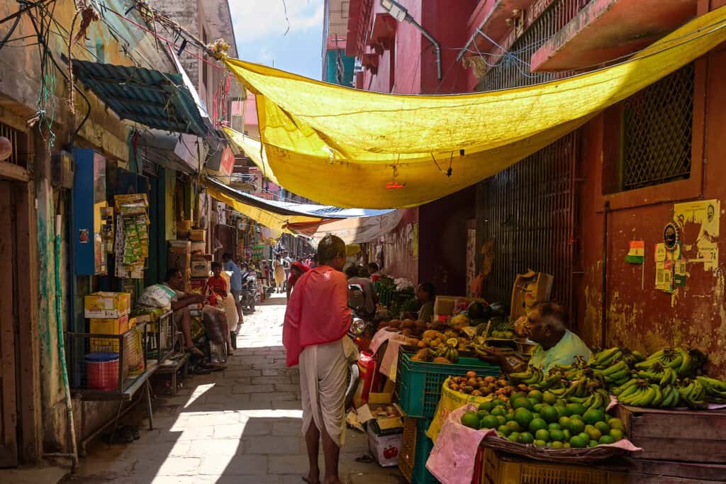 A narrow street in Varanasi with a vibrant fruit market in the foreground. Yellow tarps hang overhead, filtering bright sunlight onto the stone path. A vendor sits among crates of bananas and green citrus, while a man in a pink shawl walks away into the sun-drenched alleyway.