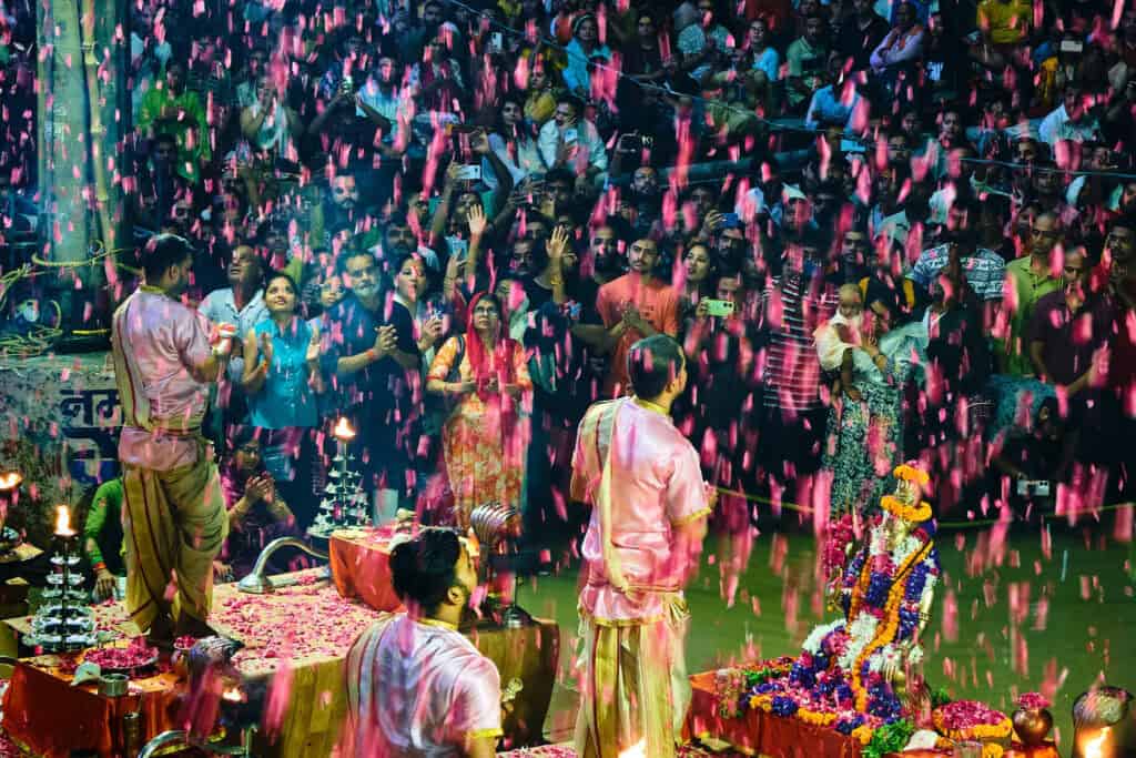 A wide, vibrant shot of the Ganga Aarti finale where bright pink flower petals rain down over the priests and the crowd. The scene is filled with motion, color, and light, capturing a moment of joyous spiritual celebration.