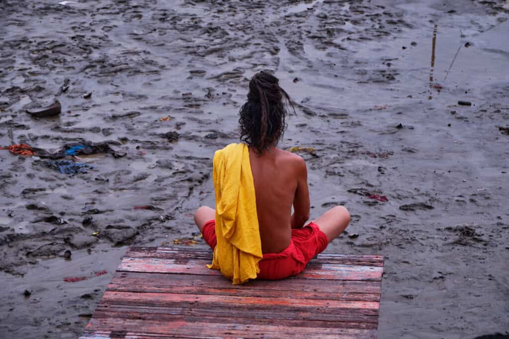 Varanasi - Seated on a weathered wooden platform, a Sadhu looks out across the mudflats, his saffron and crimson robes standing out against the muted landscape.