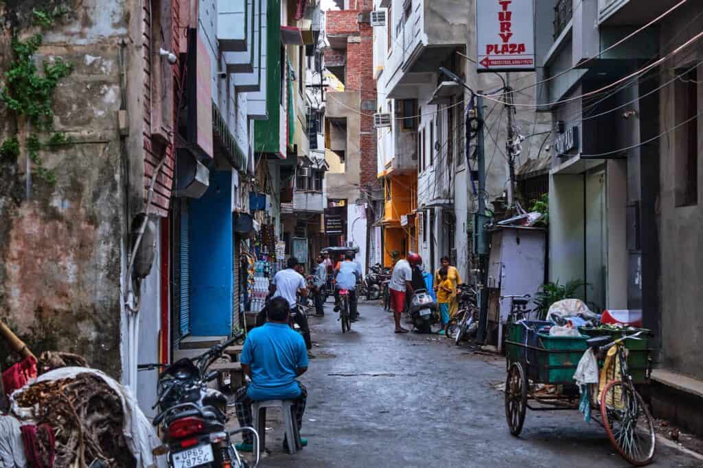 A narrow, busy street in Varanasi featuring a man in a bright blue shirt sitting on a stool in the foreground. The alleyway is lined with multi-story buildings, tangled electrical wires, and various modes of transport including motorbikes, a bicycle, and a traditional handcart.