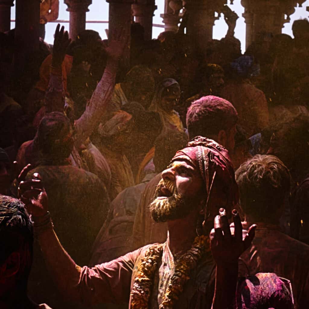 A tight, dramatic portrait of a bearded man with his face covered in yellow and red powder, looking upward with an expression of spiritual ecstasy amidst a hazy, golden background.